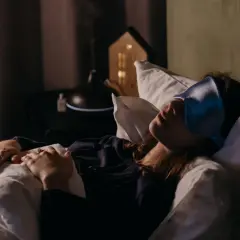 Una mujer duerme con antifaz, soñando con una persona fallecida y verla viva. A su lado, un difusor de aromas y una casita de luz.
