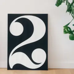 Póster minimalista con el número 2 en blanco sobre fondo negro, evocando su 2 significado espiritual. Una planta verde decora la escena.