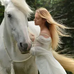 Una joven rubia con un vestido blanco se inclina hacia un majestuoso caballo blanco. Es como soñar con caballos, una escena etérea y serena.