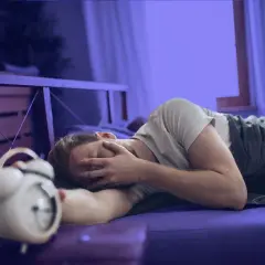 Hombre en la cama, con la cara entre las manos, junto a un despertador. Parece que me levanto cansado.