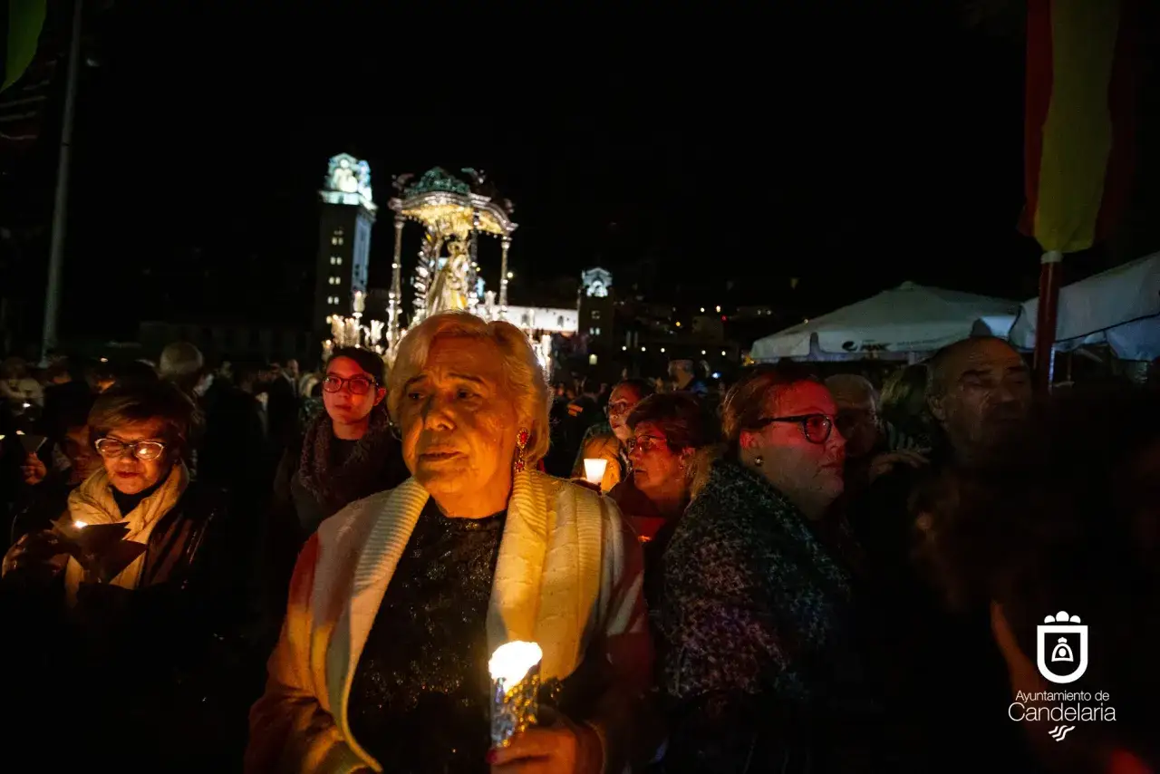 Procesión nocturna con velas. Una mujer mayor, con un chal blanco, sostiene una vela encendida. Al fondo, un paso procesional y gente. Es el santo de candela.